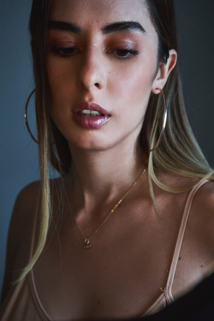 Marintia Bustos a model with a radiant complexion showcases a close-up of her face highlighting her makeup and jewelry. She wears a delicate gold necklace with a pendant and large hoop earrings and her eyes are accentuated with a warm-toned eyeshadow.